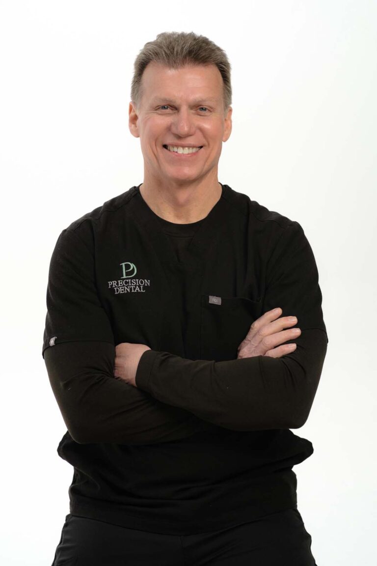 A photo of Dr. Kevin Clemmons crossing his arms and wearing a black scrubs with a logo of 'PRECISION DENTAL' on the left side of his chest, in a plain white background.