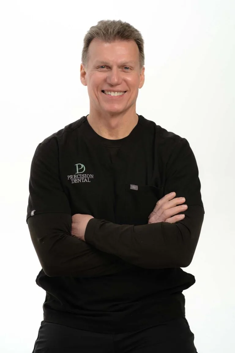 A photo of Dr. Kevin Clemmons crossing his arms and wearing a black scrubs with a logo of 'PRECISION DENTAL' on the left side of his chest, in a plain white background.
