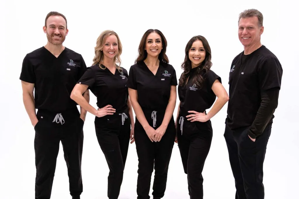 The image shows five people standing and wearing a black scrubs with a logo of 'PRECISON DENTAL'. The background is plain white.